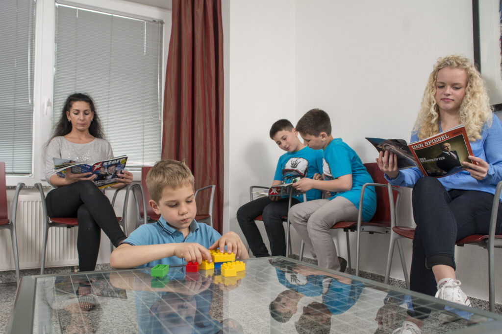 Wartezimmer mit einer Frau, zwei Jungen und einem Mädchen am Lesen. Ein Kind spielt auf einem Tisch mit Bauklötzen.